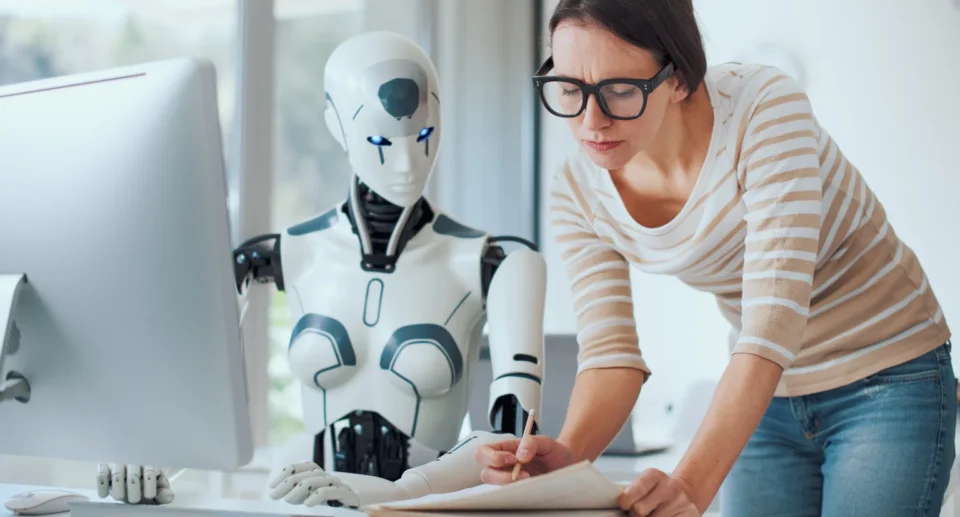 A woman working alongside a humanoid robot at a desk, reviewing notes while the robot interacts with a desktop computer.