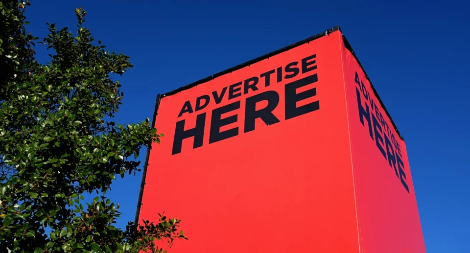 Large red billboard with the text “ADVERTISE HERE” in black capital letters, set against a clear blue sky and partially framed by green leaves.