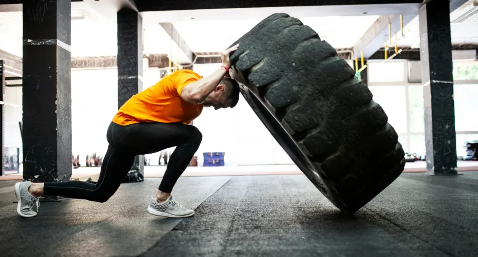 A man in an orange shirt and black leggings pushes a massive tire upward in a gym, engaging in a full-body strength workout.