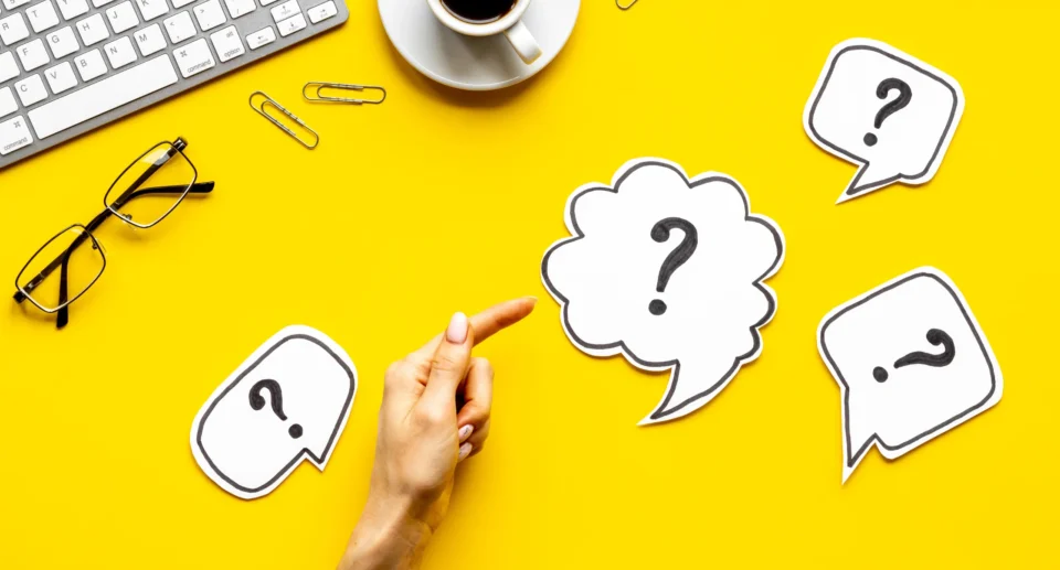 A hand pointing at paper cut-out question marks on a yellow desk with a keyboard, glasses, and coffee, symbolizing FAQ creation and strategy